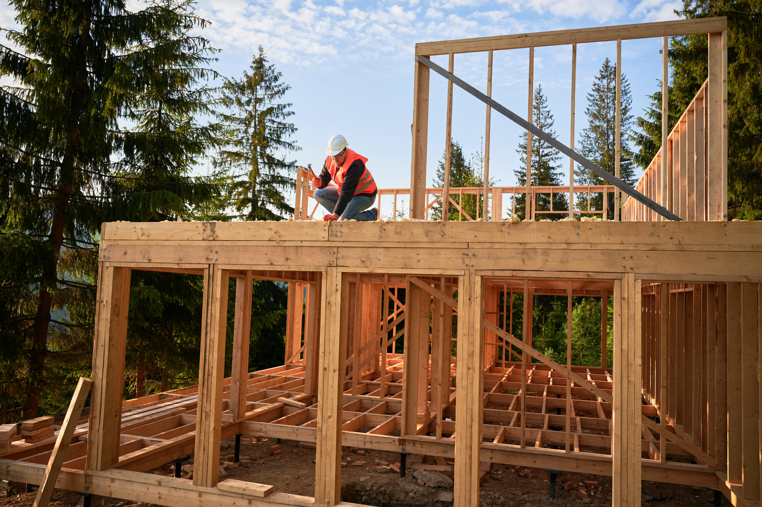 Carpenter constructing two-story wooden frame house near forest. Bearded man hammering nails into the structure, wearing protective helmet, construction vest. Concept of modern ecological construction